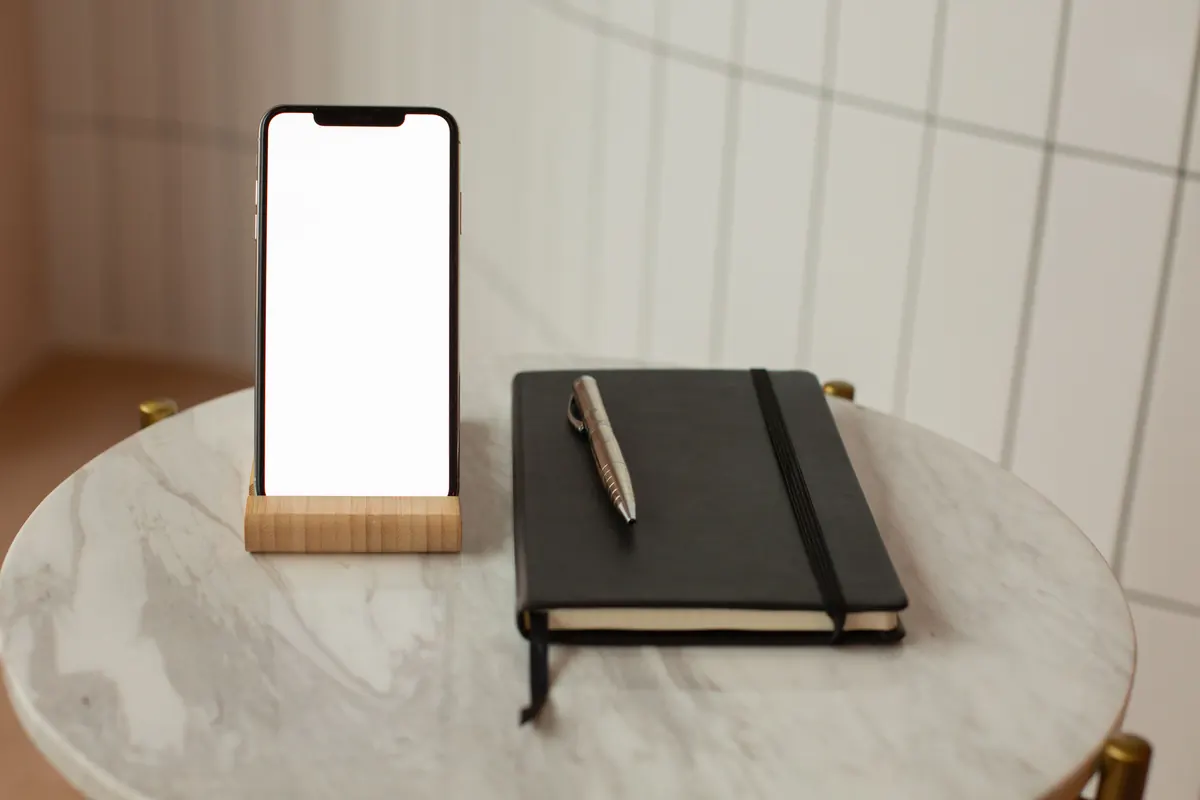 Blank-screen phone on a wooden stand beside a notebook and pen.