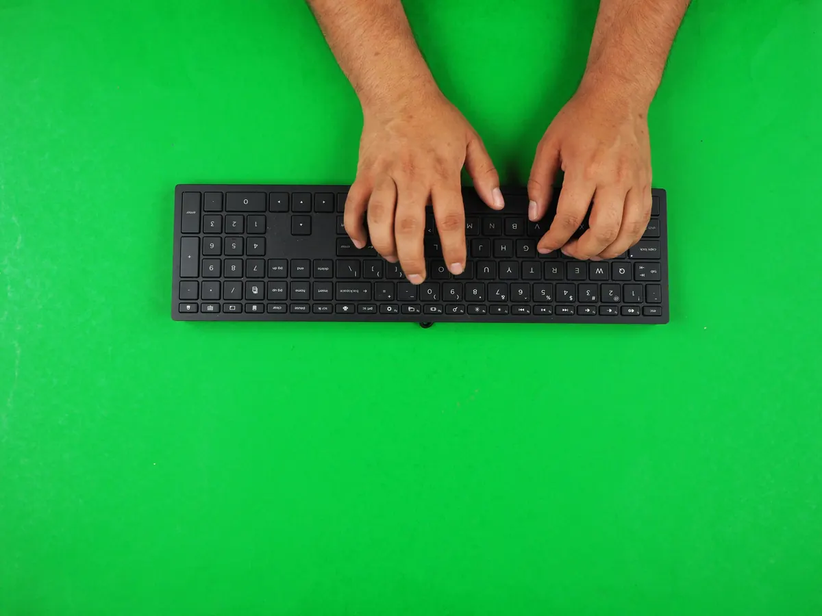 Two hands typing on a plain black full-size keyboard on a green surface, seen from above.