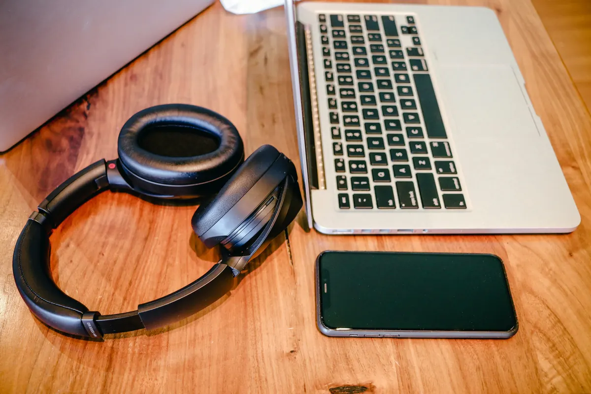 Wireless over-ear headphones, an open laptop and a smartphone with a dark screen on a wooden desk.