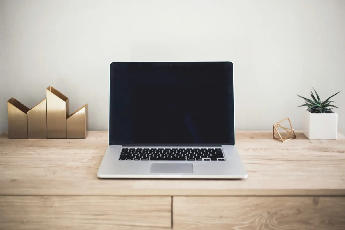 An open laptop with a dark screen on a light wooden desk between small decorative objects and a plant.