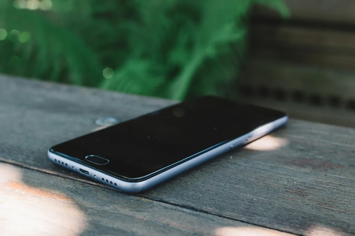 A smartphone with its screen off lying on a weathered wooden bench, with green leaves behind it.