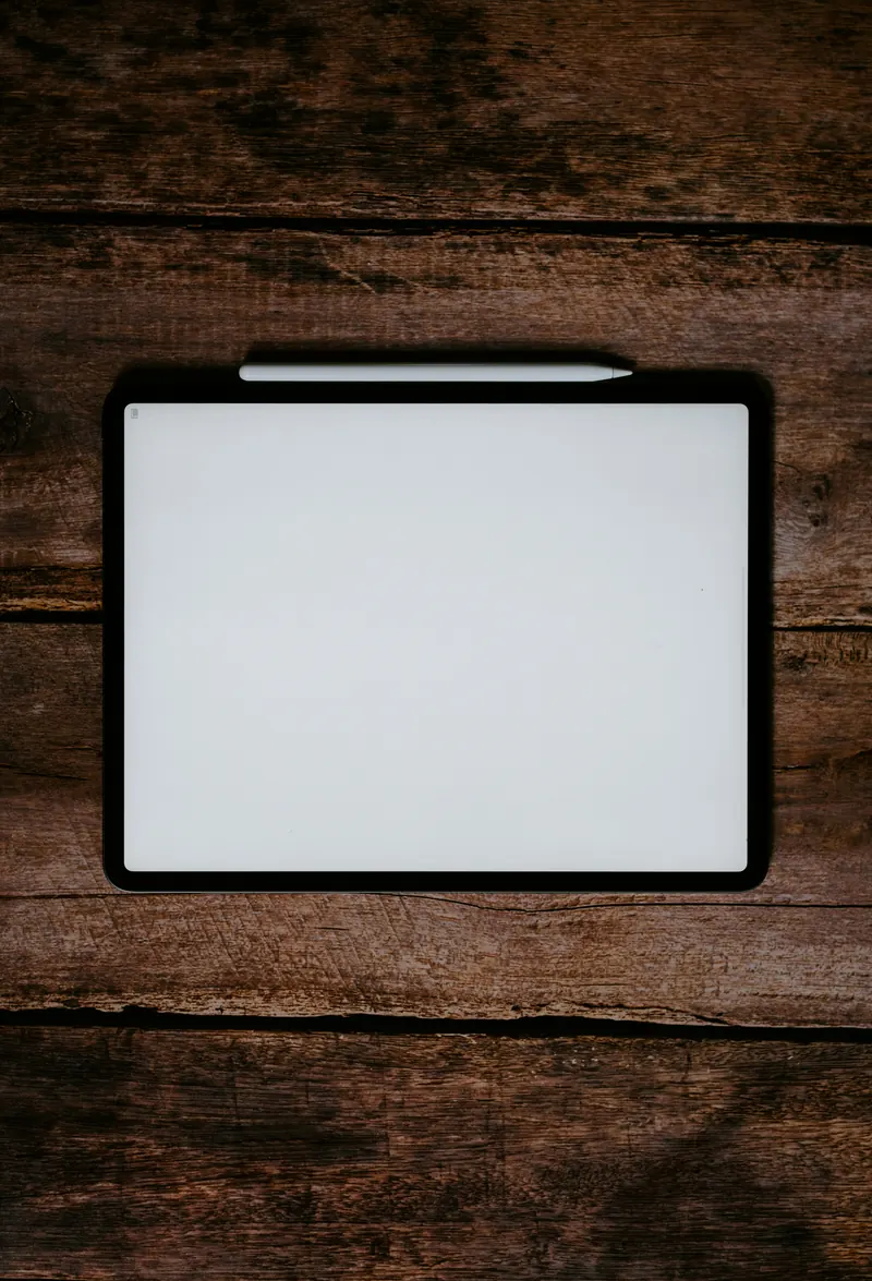 A tablet with a blank white screen and a stylus, seen from above on a dark wooden table.