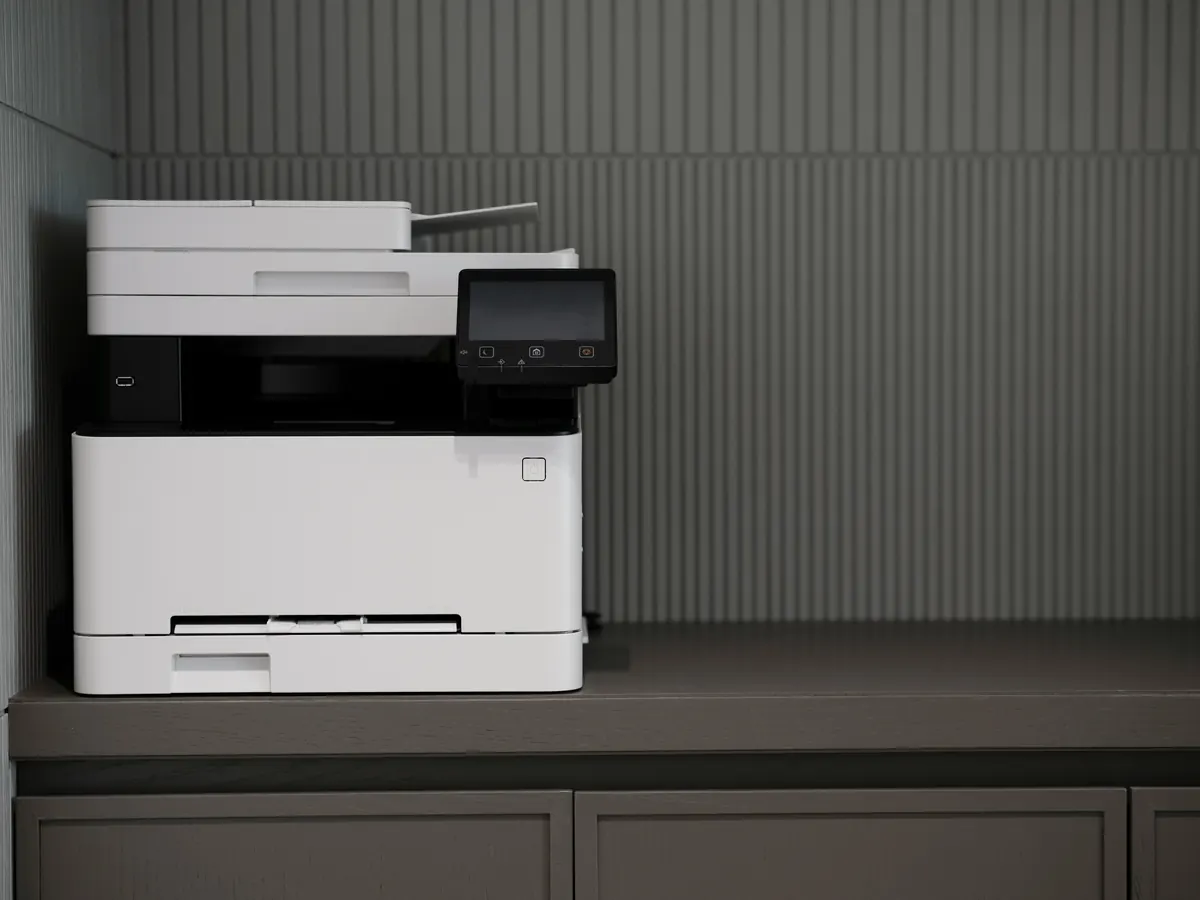 A white multifunction printer with its control screen off, standing on a gray counter against a ribbed wall.